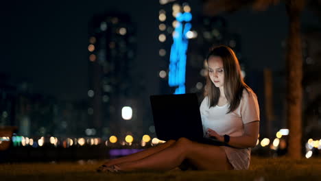 A-young-woman-on-a-summer-night-sitting-on-the-grass-in-the-city-looks-at-the-laptop-screen-and-writes-a-message-on-the-keyboard.-Programmer-freelancer.-Remote-night-work